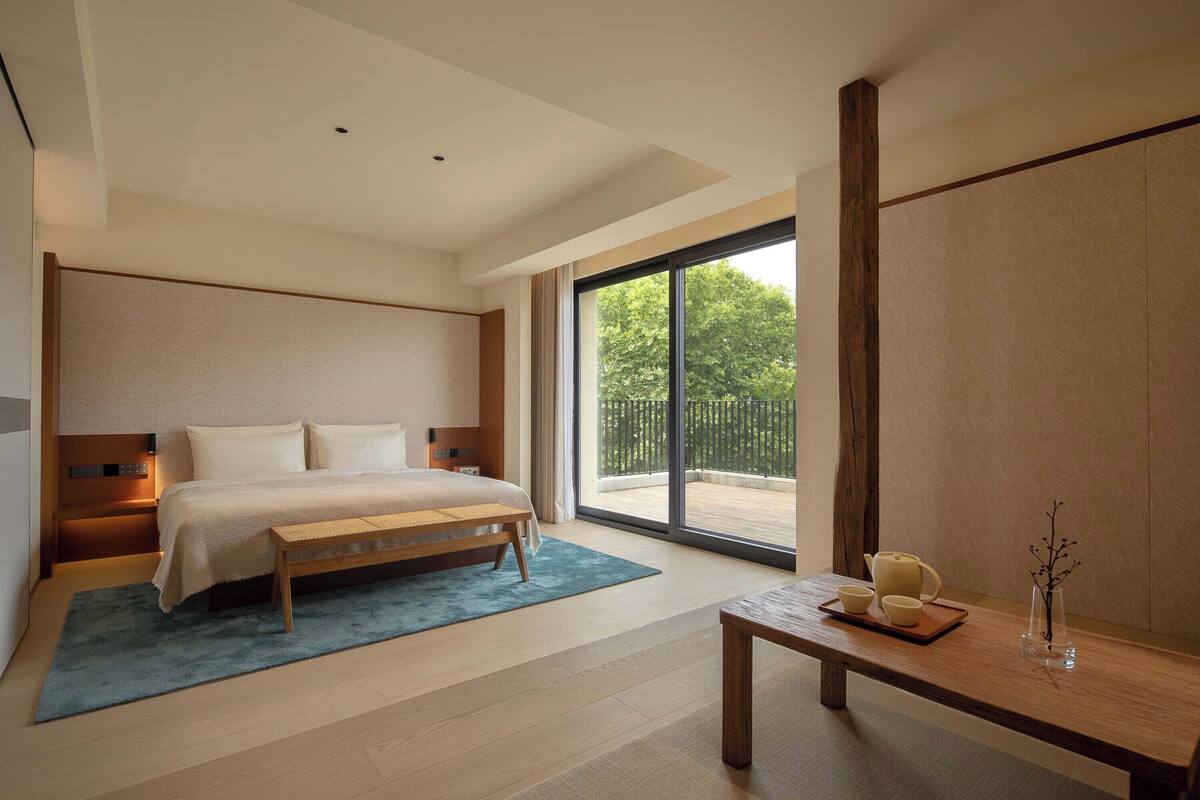 Un dormitorio moderno con una cama grande, un banco de madera, una pequeña mesa con juego de té y vistas a la vegetación por la ventana.