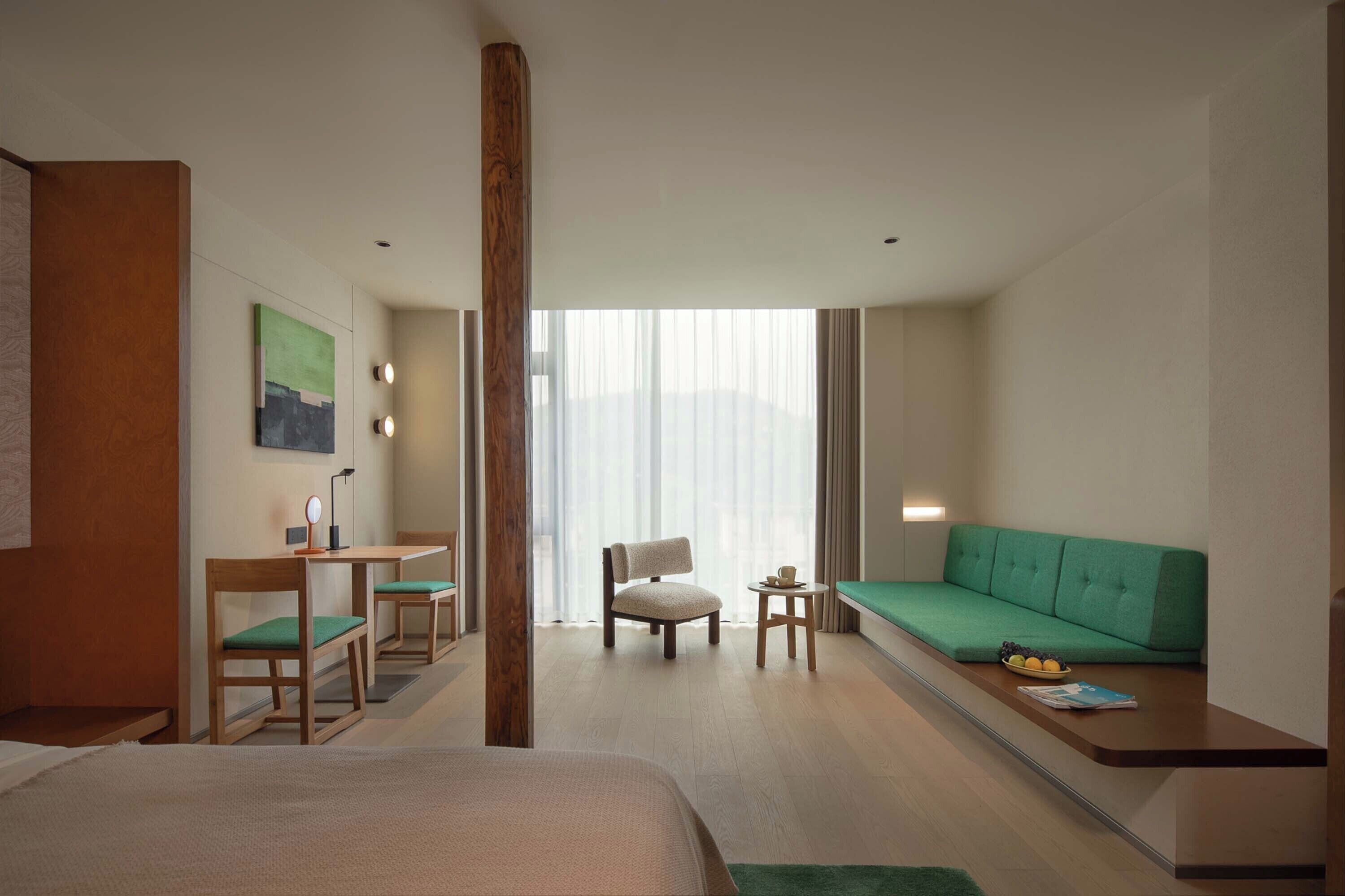 Une chambre d’hôtel avec un lit, un bureau et une chaise, un canapé vert et une vue sur l’extérieur grâce à de grandes fenêtres.