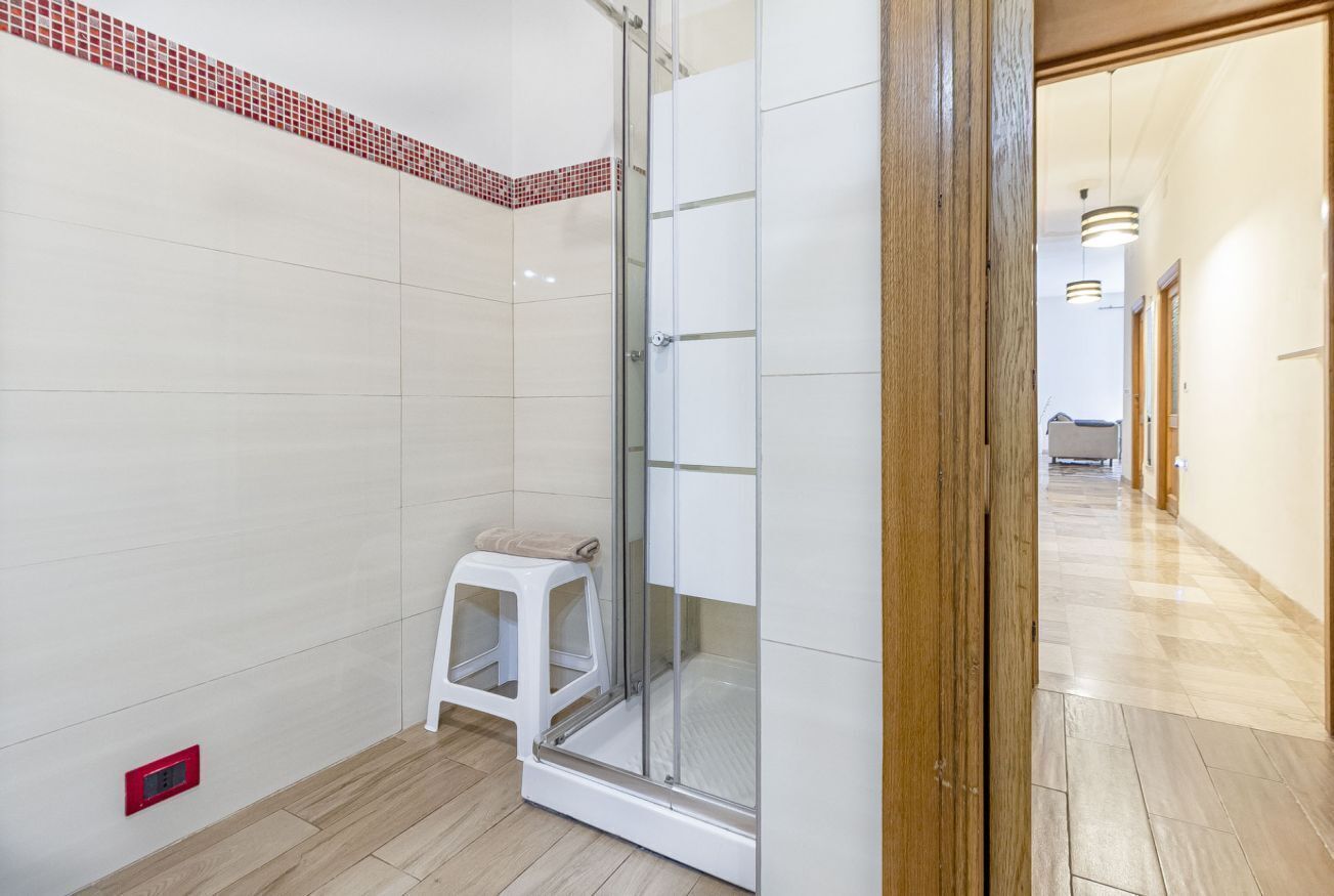 A modern bathroom with a glass shower enclosure, a white stool, and a red electrical outlet on the floor.