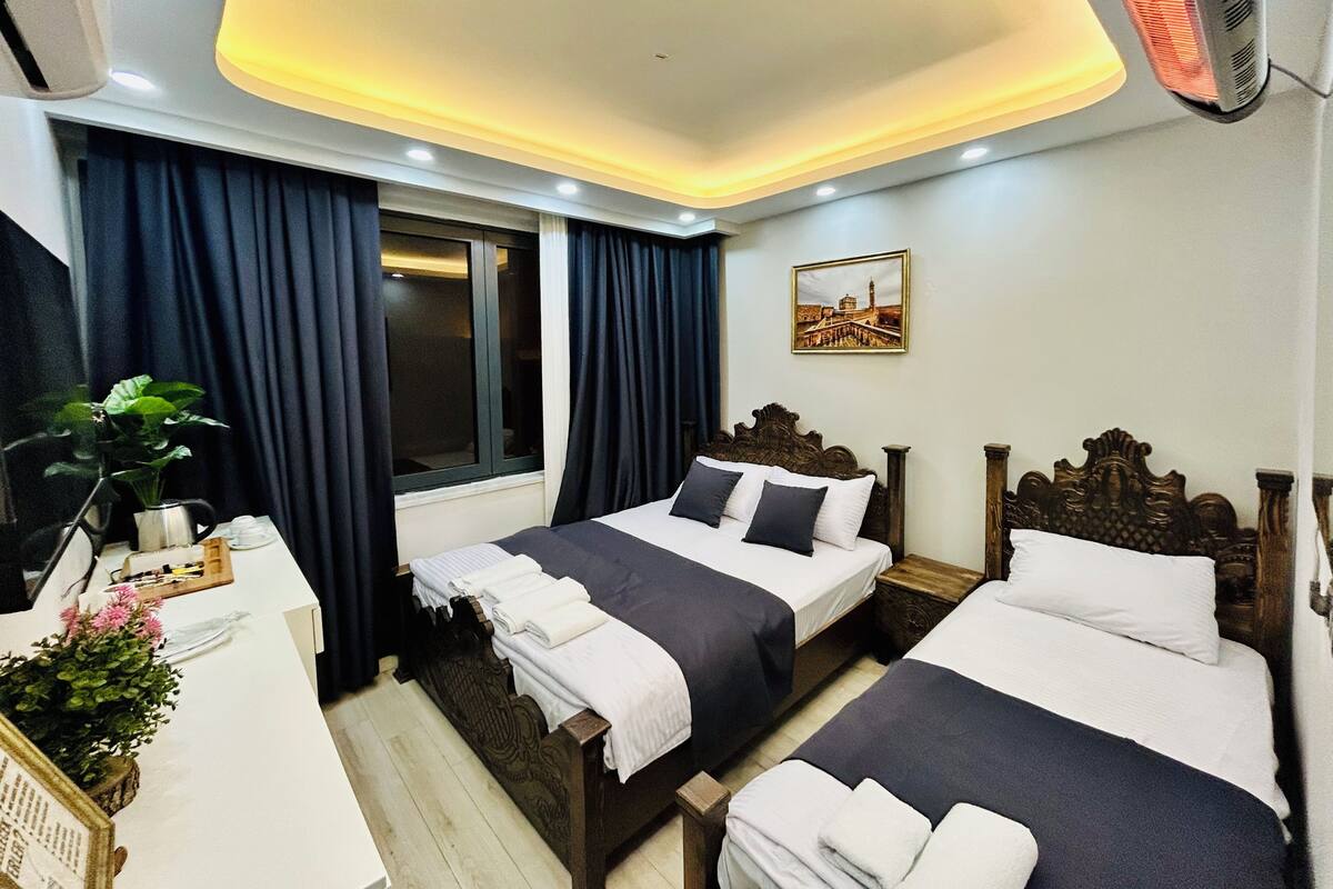 Une chambre d’hôtel avec deux lits, un bureau, une chaise, une fenêtre avec des rideaux, un radiateur fixé au mur et un tableau encadré.