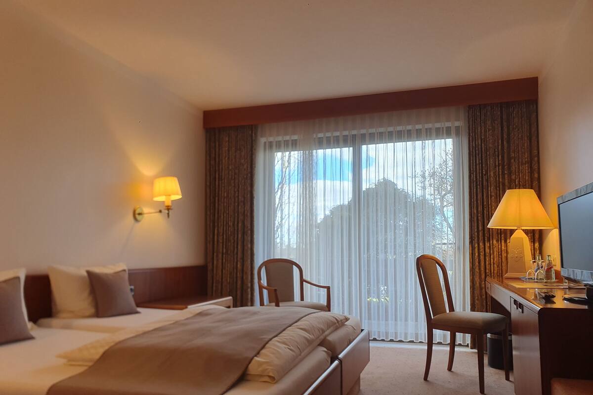 A hotel room with a bed, two chairs, a desk with a lamp, and a TV.