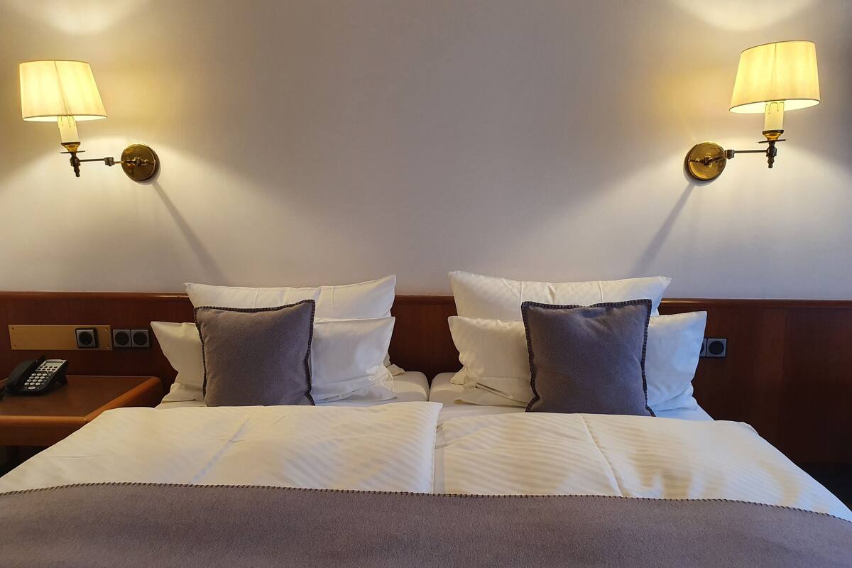 A neatly made bed with white and gray bedding, two wall-mounted lamps, a bedside phone, and a wooden headboard.