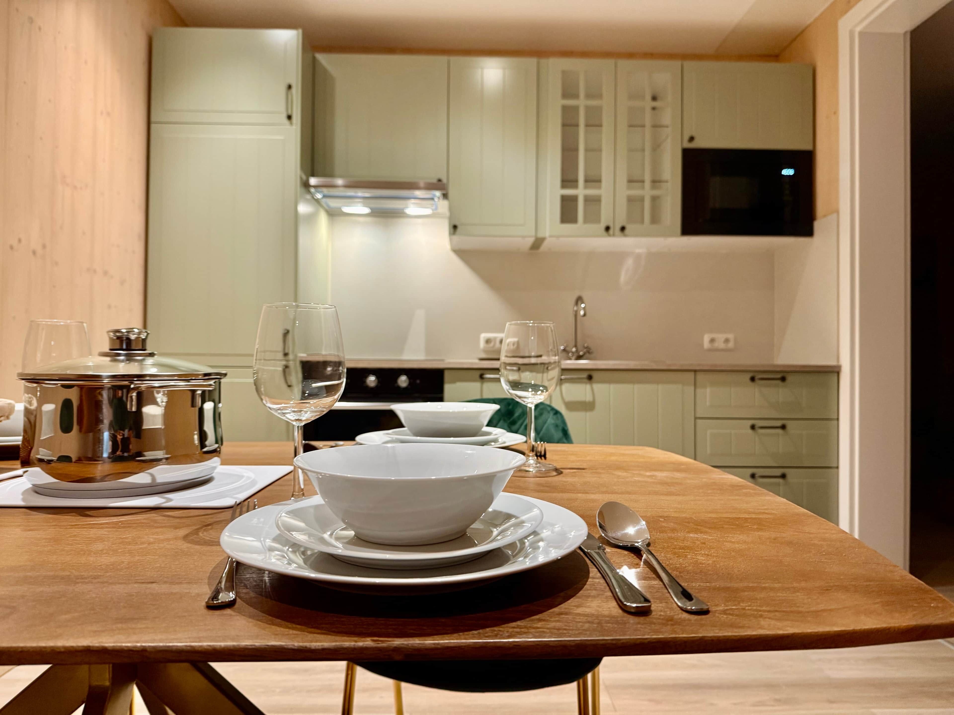 A dining table set with white plates, bowls, and cutlery in a modern kitchen with wooden cabinets and a microwave.