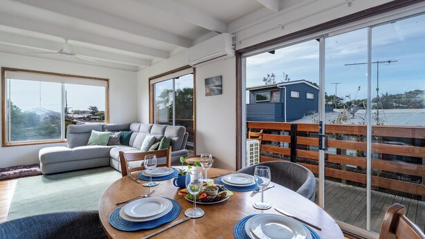 A dining area with a wooden table set for four, a gray sofa, and a large window with a view of a blue house and trees. - Walk to Beach | Pet Friendly | Sleep 5 (Ocean Grove)