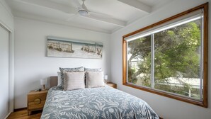 A bedroom with a large bed, a nightstand, a ceiling fan, a window with a view of trees, and a beach-themed painting on the wall. - Walk to Beach | Pet Friendly | Sleep 5 (Ocean Grove)