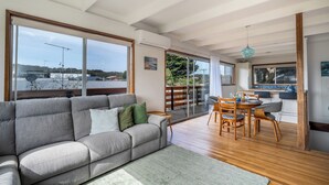 A spacious living room with a grey sofa, wooden dining table, and large windows offering a view of the outside. - Walk to Beach | Pet Friendly | Sleep 5 (Ocean Grove)