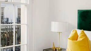 A hotel room with a large window, a bed with a patterned bedspread, a nightstand with a lamp, and a green accent wall. - Chic Beauchamp Place Home (London)