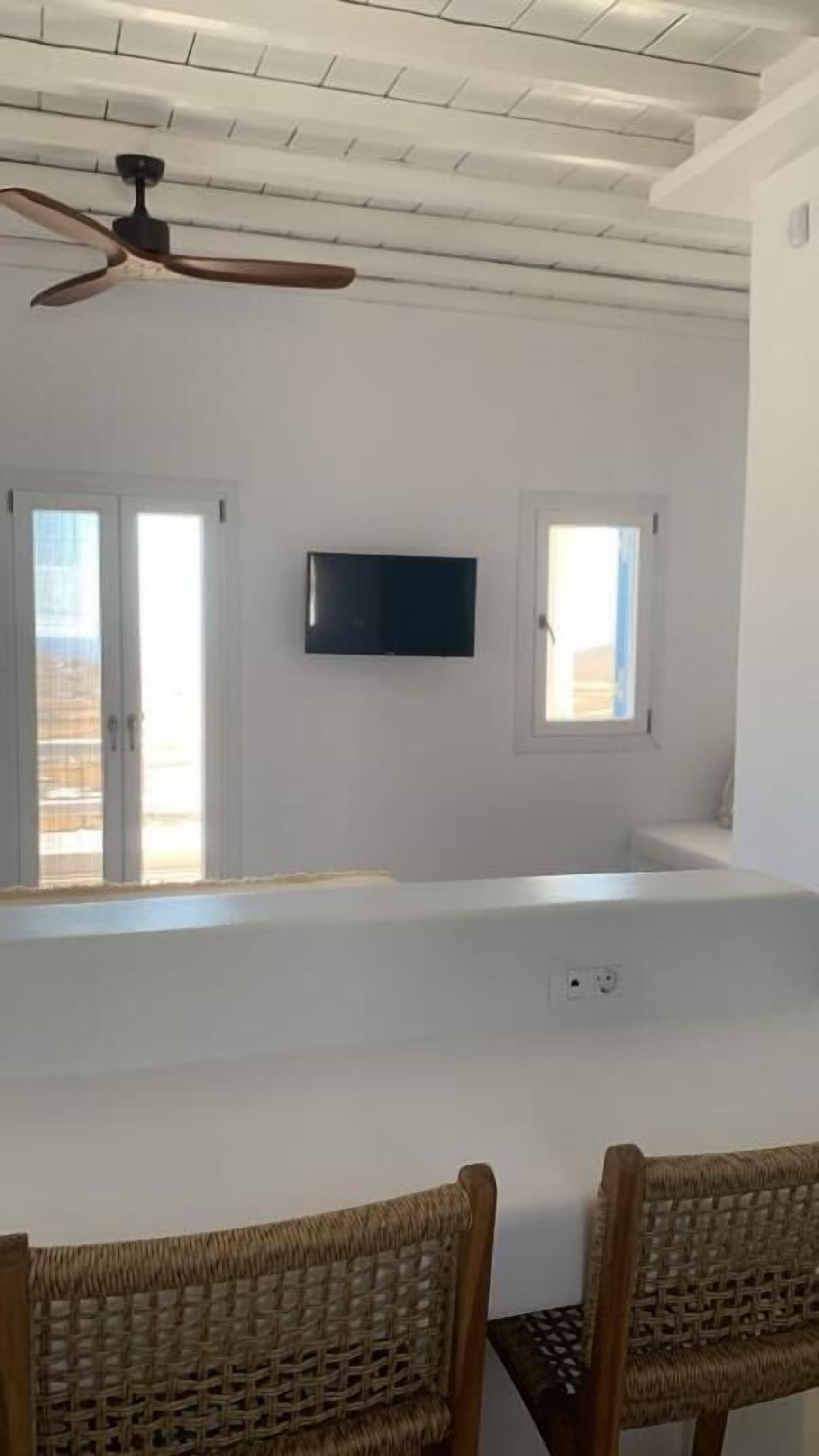 Ένα δωμάτιο με έναν ανεμιστήρα οροφής, μια τηλεόραση επίπεδης οθόνης τοποθετημένη στον τοίχο και δύο παράθυρα με λευκά πλαίσια.