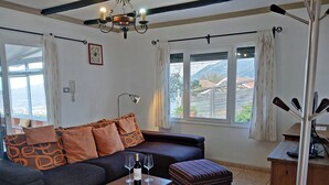 Ein Wohnzimmer mit einem dunklen Sofa, gemusterten Kissen, einem kleinen Holztisch mit Weingläsern, einem gestreiften Koffer und Blick auf eine gebirgige Landschaft durch große Fenster.