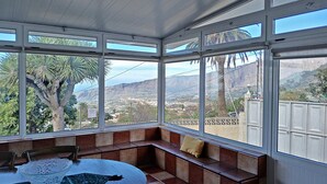 Ein Wintergarten mit großem Fenster und Ausblick auf eine reizvolle Landschaft, ein runder Tisch mit Glasvase und eine Bank mit Kissen.