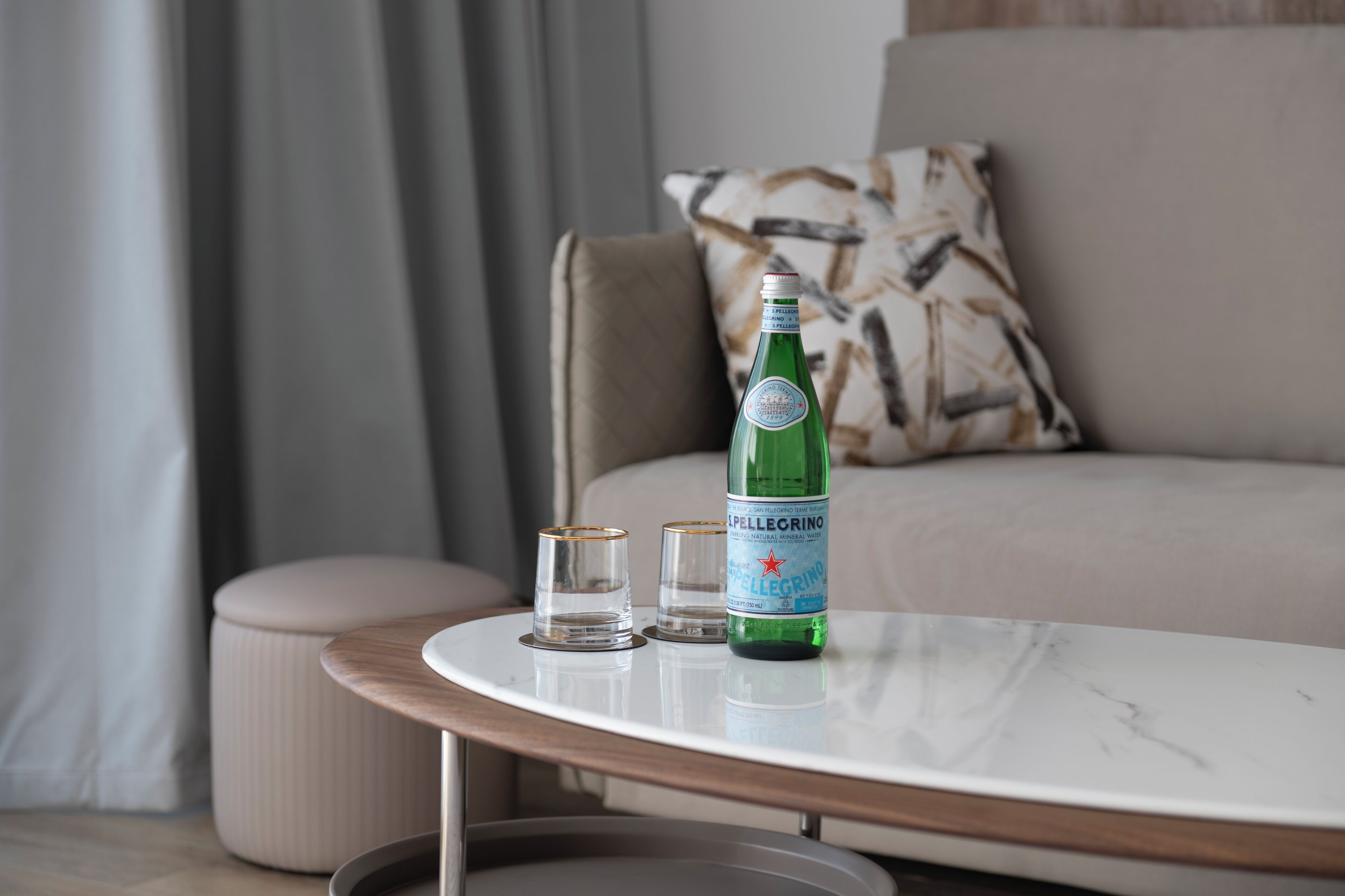 En grön flaska Pellegrino-vatten, två glas och ett bord i marmor i ett modernt vardagsrum med en soffa och en pall.