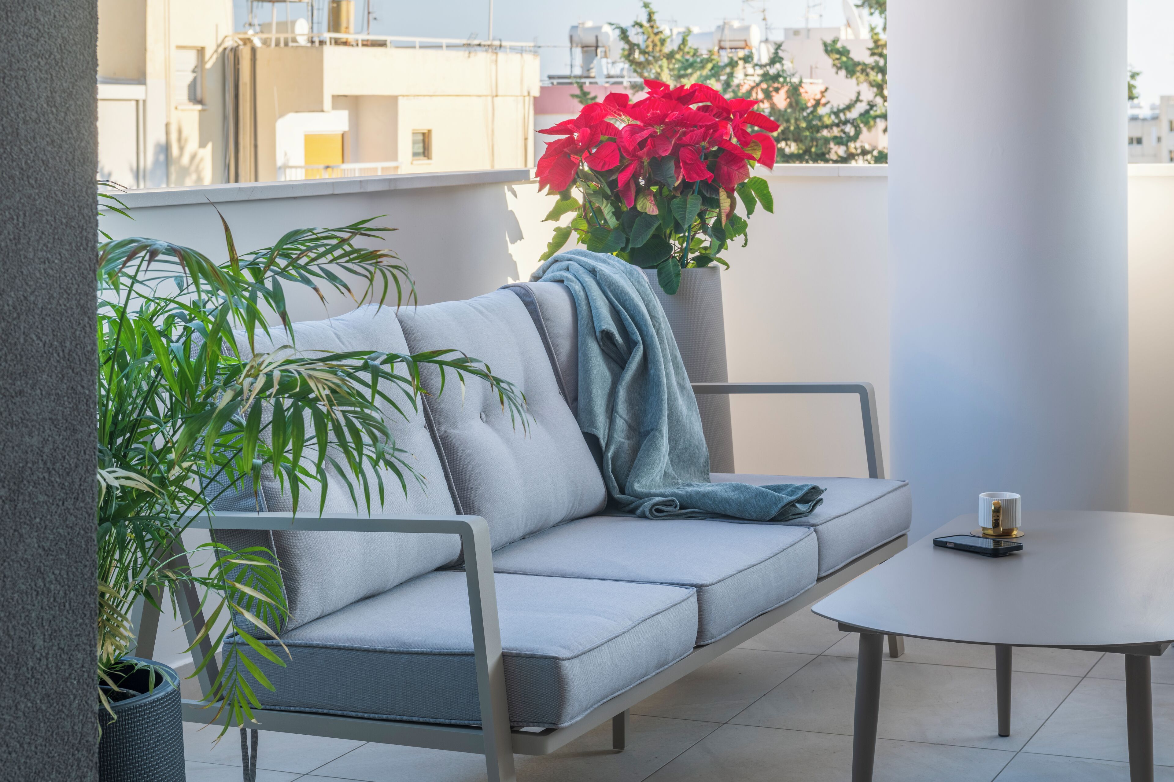 Een balkon met een grijze bank, een klein tafeltje en een potplant met rode bloemen.