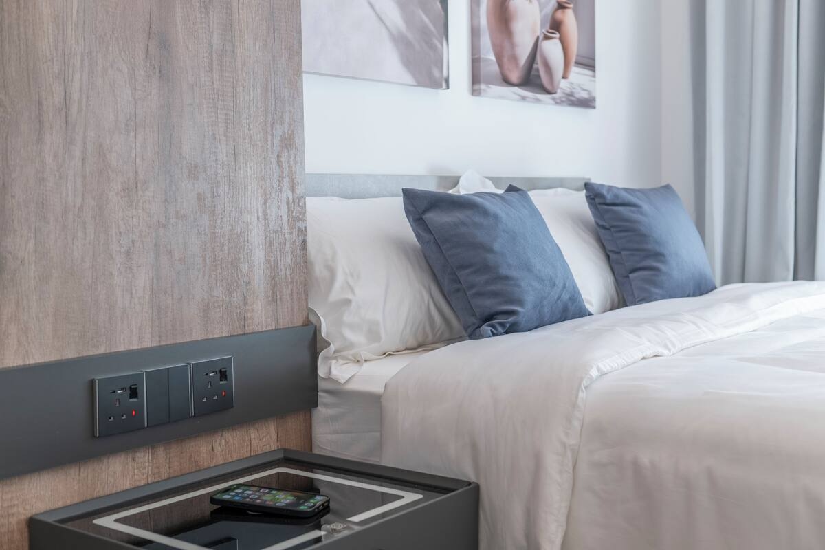 Una camera d'hotel con una testiera in legno, un letto con biancheria da letto bianca e blu, un comodino con un telecomando e una presa di corrente a parete.