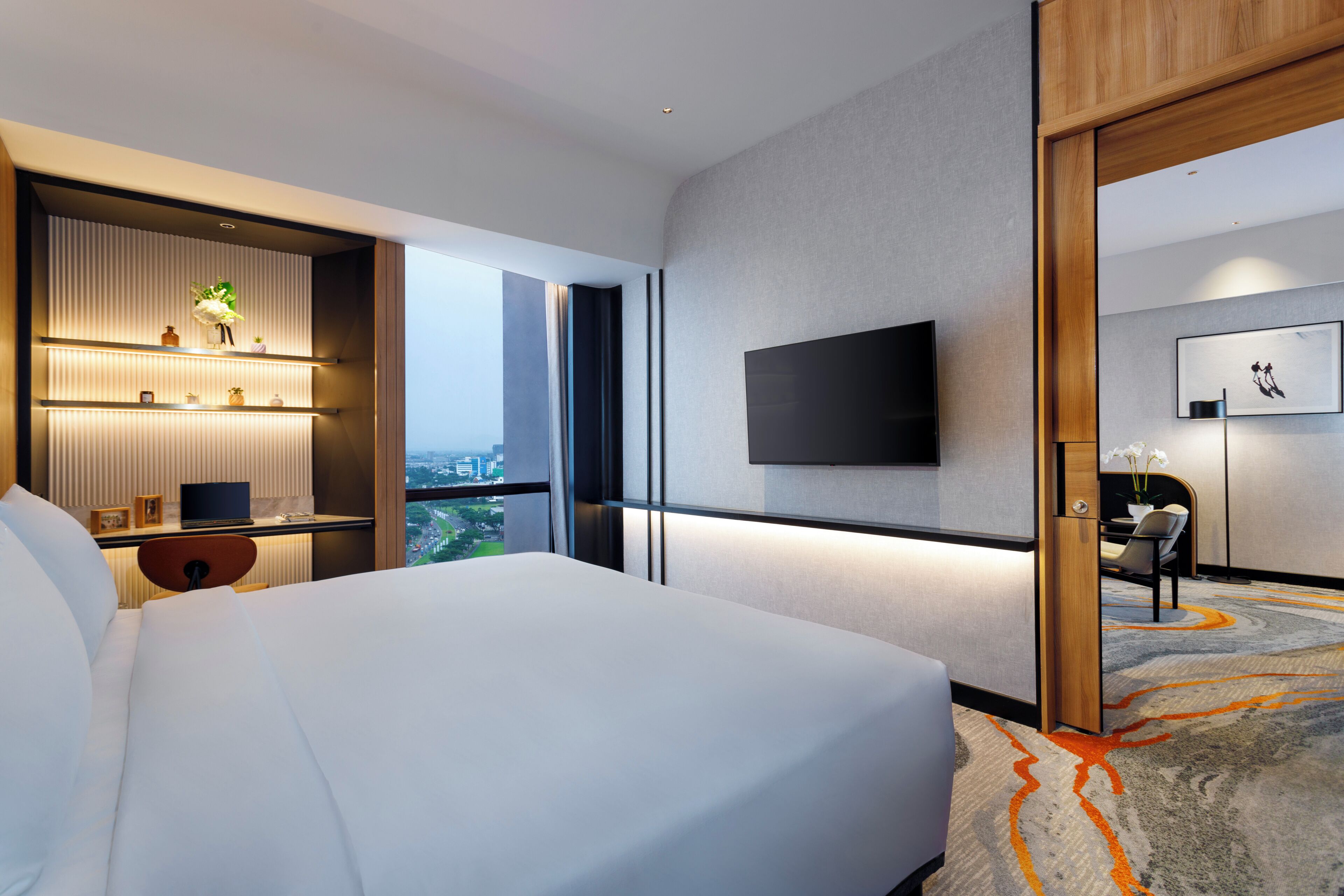 A modern hotel room with a large bed, a desk with a computer, a flat-screen TV mounted on the wall, and a view of the cityscape.