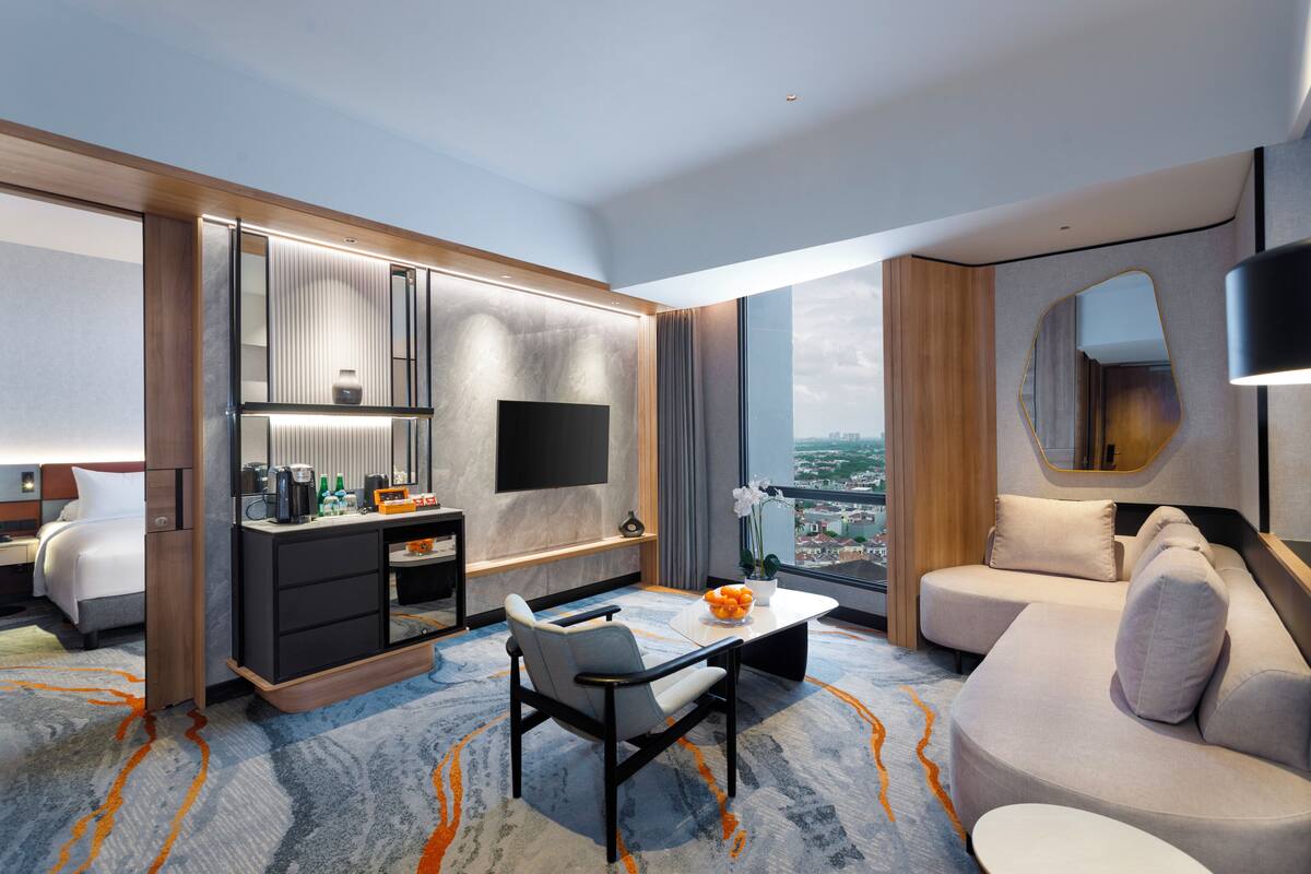 A modern hotel room with a bed, a desk with a flat-screen TV, a sofa, and a large window offering a city view.