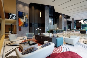 A modern hotel lobby with a large seating area, a reception desk, and a view of the kitchen.