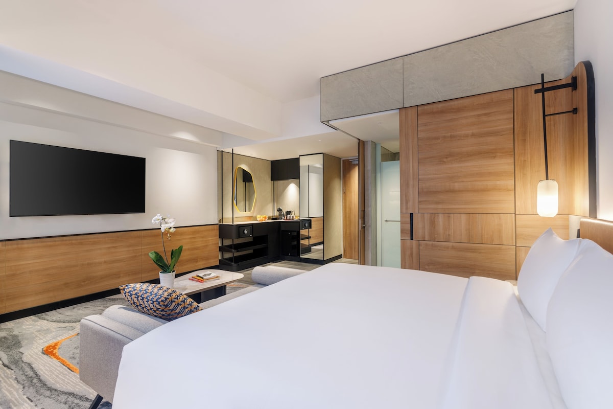 A modern hotel room with a large bed, a flat-screen TV mounted on the wall, a small seating area with a sofa and a coffee table, and a separate kitchenette area.