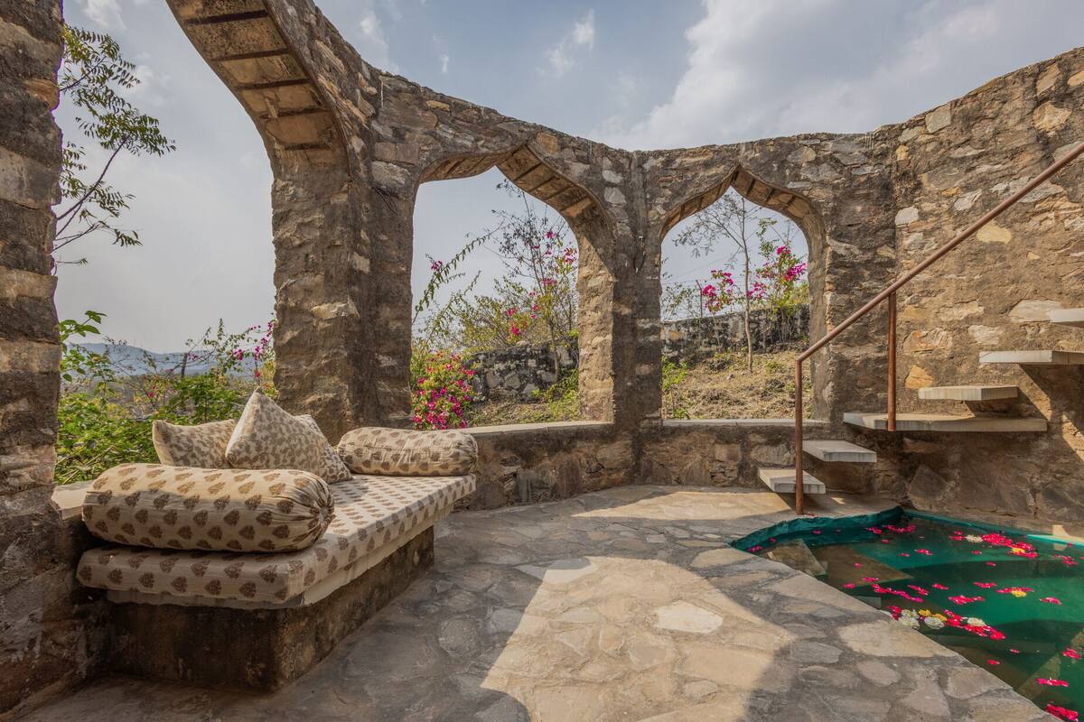 A stone-walled room with a stone bench and cushions, a small pool with floating flowers, and a staircase.
