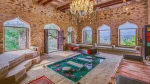 A stone-walled room with a large chandelier, a pool with floating platforms, and multiple windows offering a view of greenery. - Raht Manmatha (Udaipur)