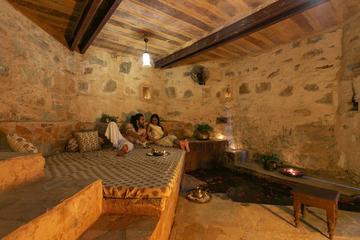A stone-walled room with a low ceiling, a bed with patterned bedding, a small table with a candle, and a bench.