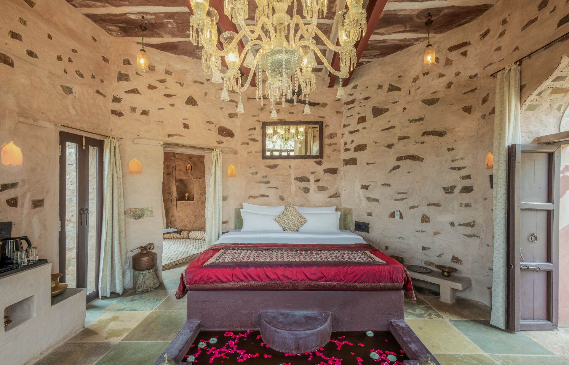 Un dormitorio con pared de piedra, un candelabro, una cama con colcha roja y una pequeña fuente decorada con flores.