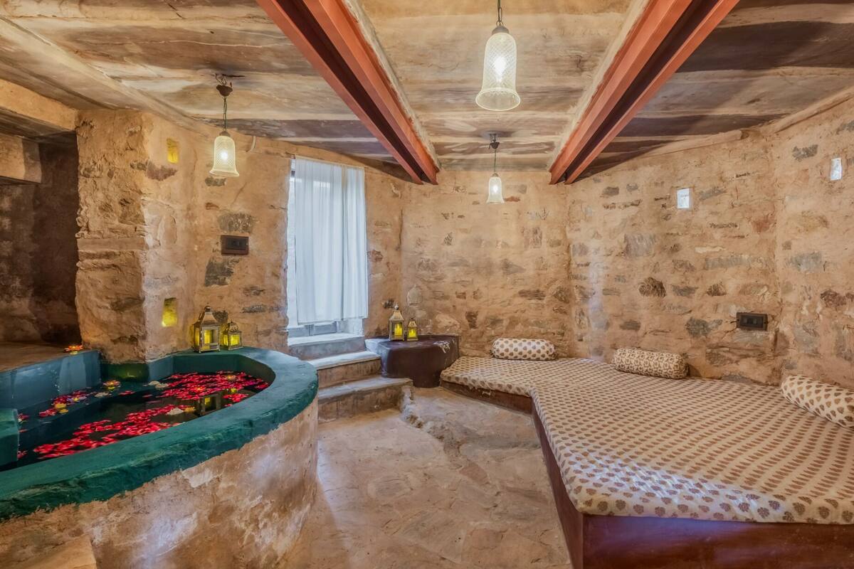 A rustic room with stone walls, a bed with patterned bedding, a green bathtub with red flowers, and hanging light fixtures.