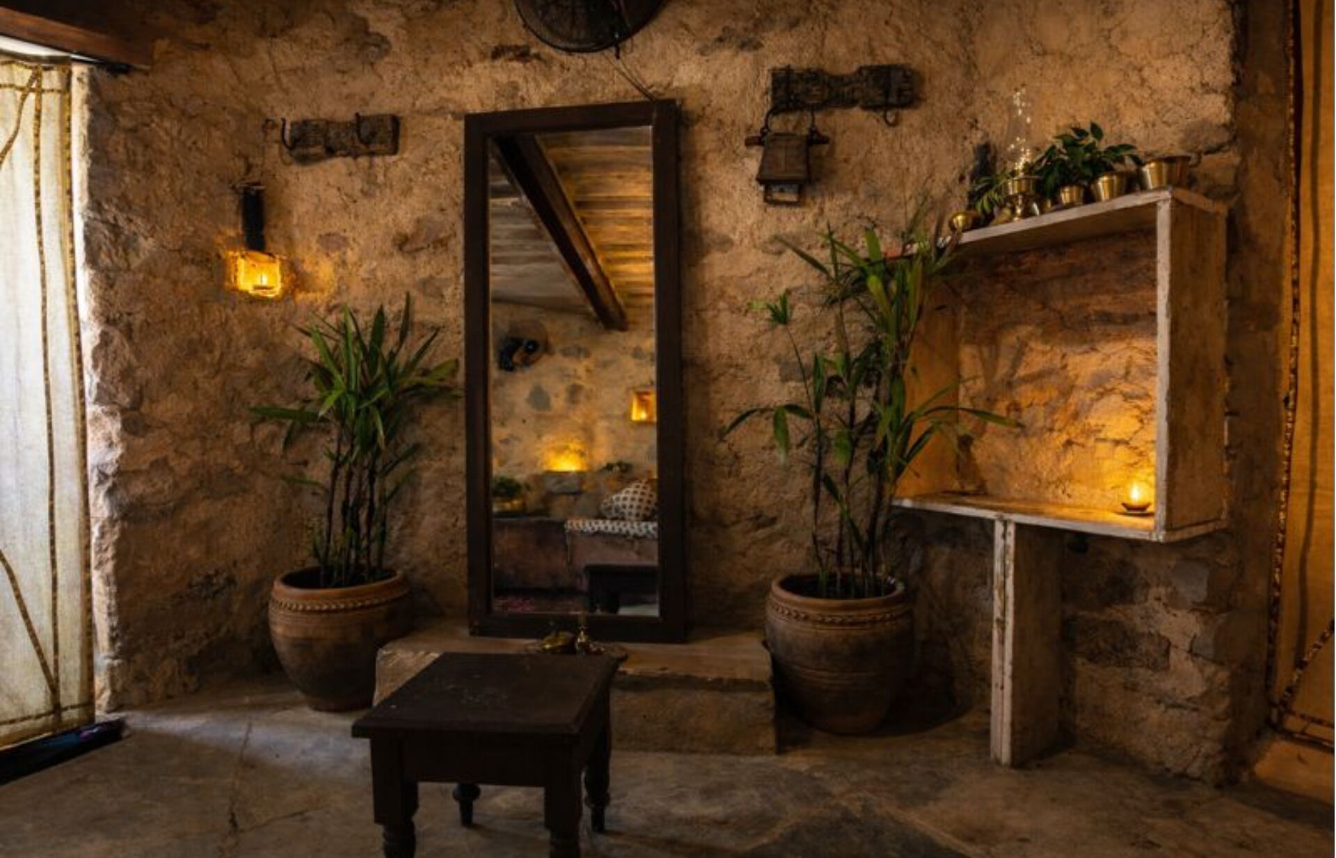 A rustic interior with stone walls, a wooden table, potted plants, and a mirror.