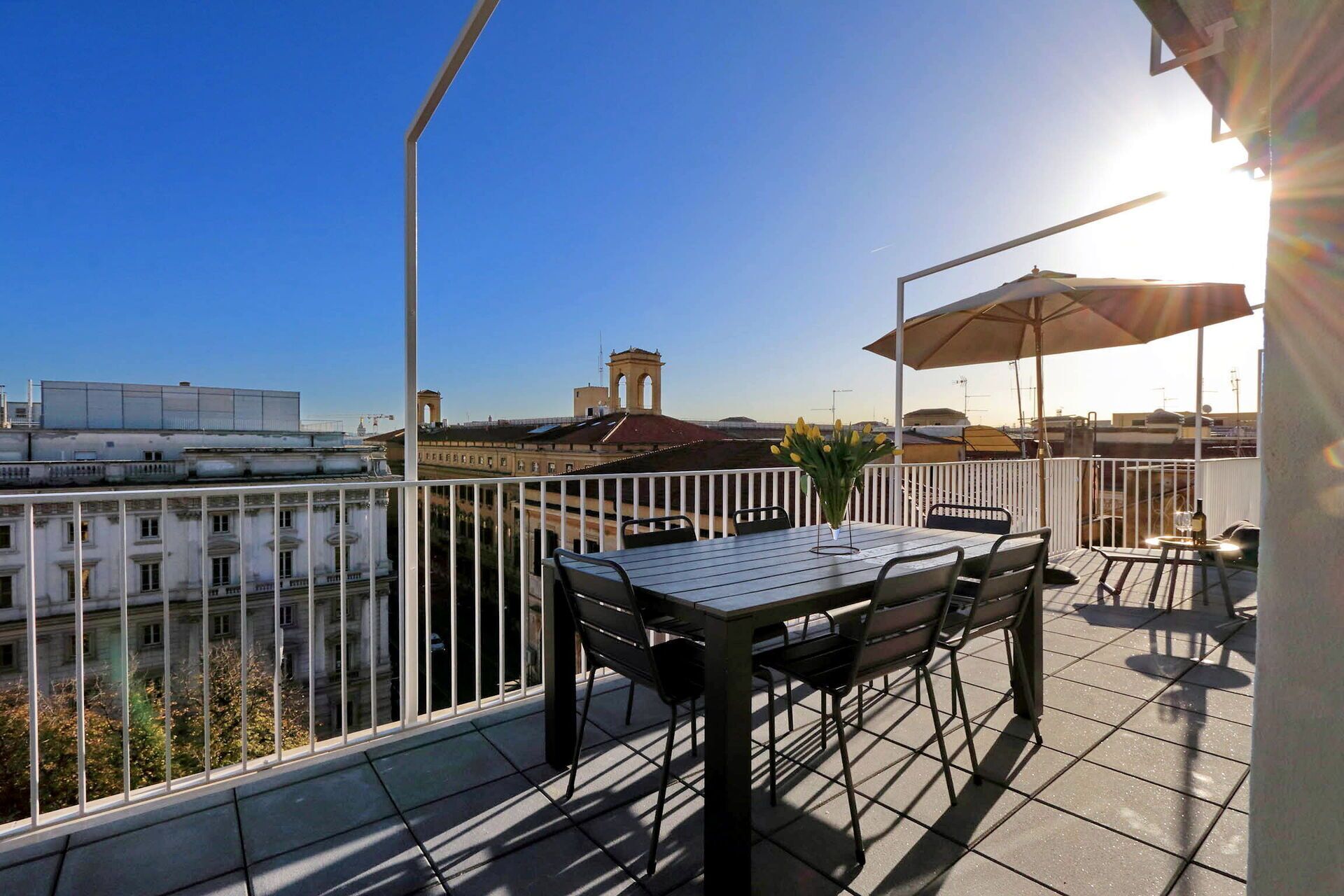 A balcony with a table and chairs, a vase of flowers, and a view of buildings and a tower.