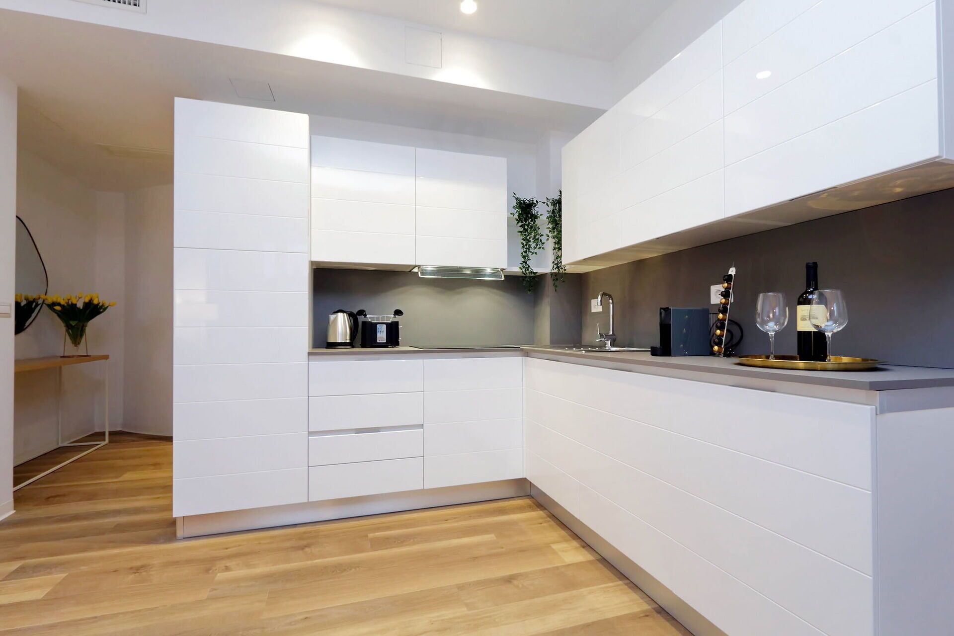 Une cuisine moderne avec des meubles blancs, un plancher en bois et un meuble à vin contenant des bouteilles et des verres.