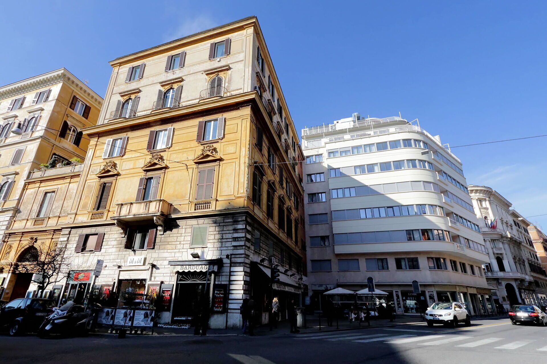 Vista de la calle con un edificio histórico que tiene una tienda en la planta baja y un edificio moderno contiguo con varios pisos y grandes ventanales.