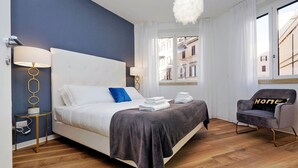 A modern hotel room with a large bed, two bedside lamps, a grey armchair with a 'Home' pillow, and a view of the city through the window. - FlaviaDesign City Suites Rome by Halldis (Rome)