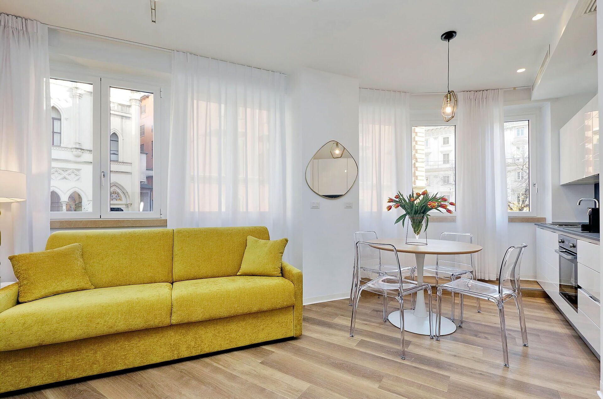 A bright living room with a yellow sofa, a round dining table with chairs, and a kitchen area with modern appliances.
