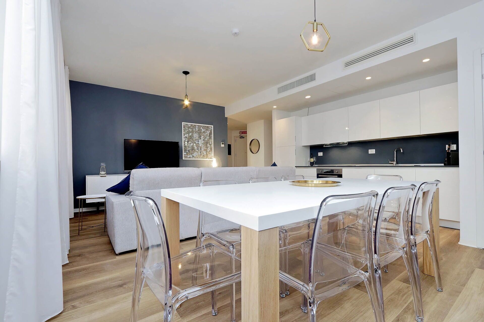 Une cuisine moderne avec des meubles blancs, une table à manger entourée de chaises transparentes et un téléviseur en arrière-plan.