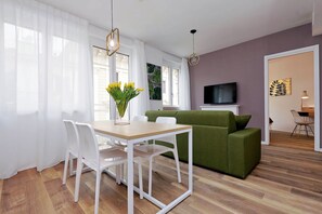 Moderni olohuone, jossa on vihreä sohva, valkoiset tuolit, puinen ruokapöytä ja seinälle asennettu televisio.