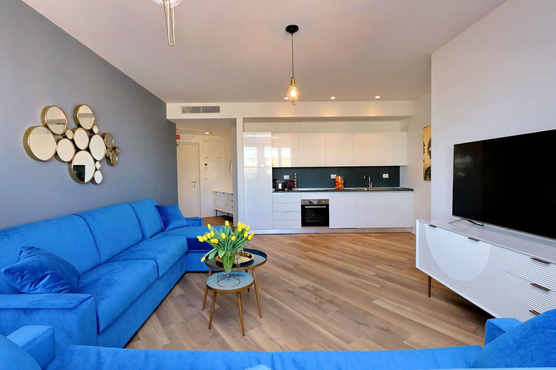 A modern living room with a blue sofa, a small round table with a vase of flowers, a flat-screen TV, and a kitchen area with white cabinets and a black countertop.