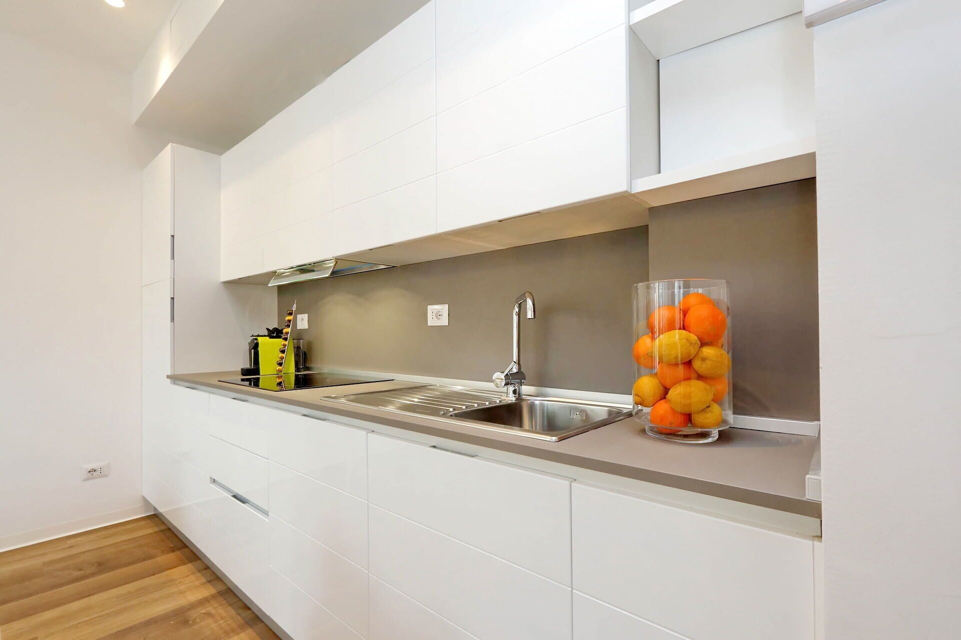 Une cuisine moderne avec des meubles blancs, un évier en acier inoxydable et un récipient en verre contenant des oranges.
