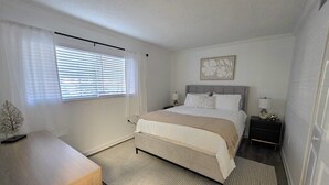 Un dormitorio con cama, mesitas de noche, lámpara, ventana con persianas y una obra de arte en la pared.