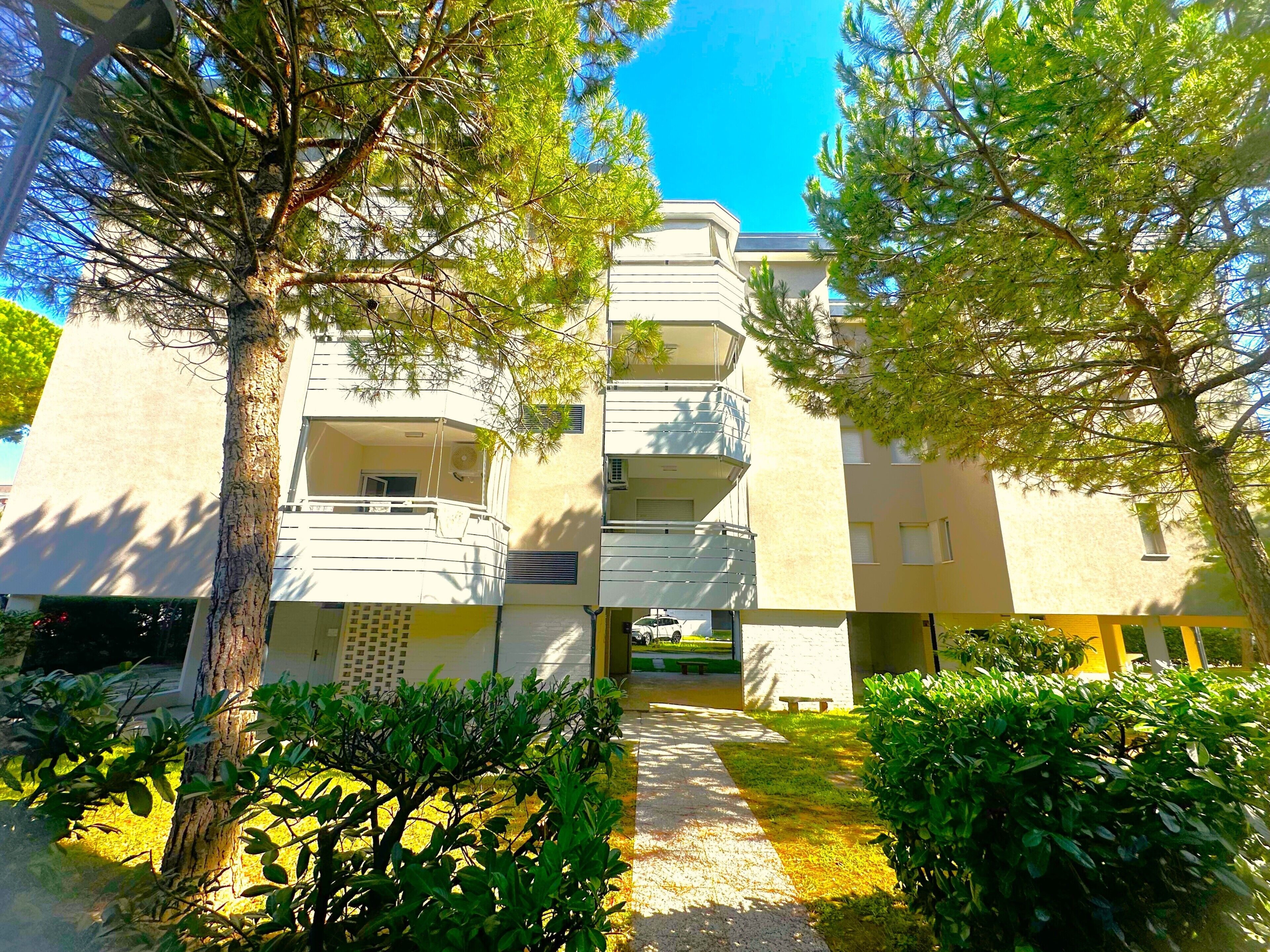 A multi-story building with balconies, surrounded by greenery and a clear sky.