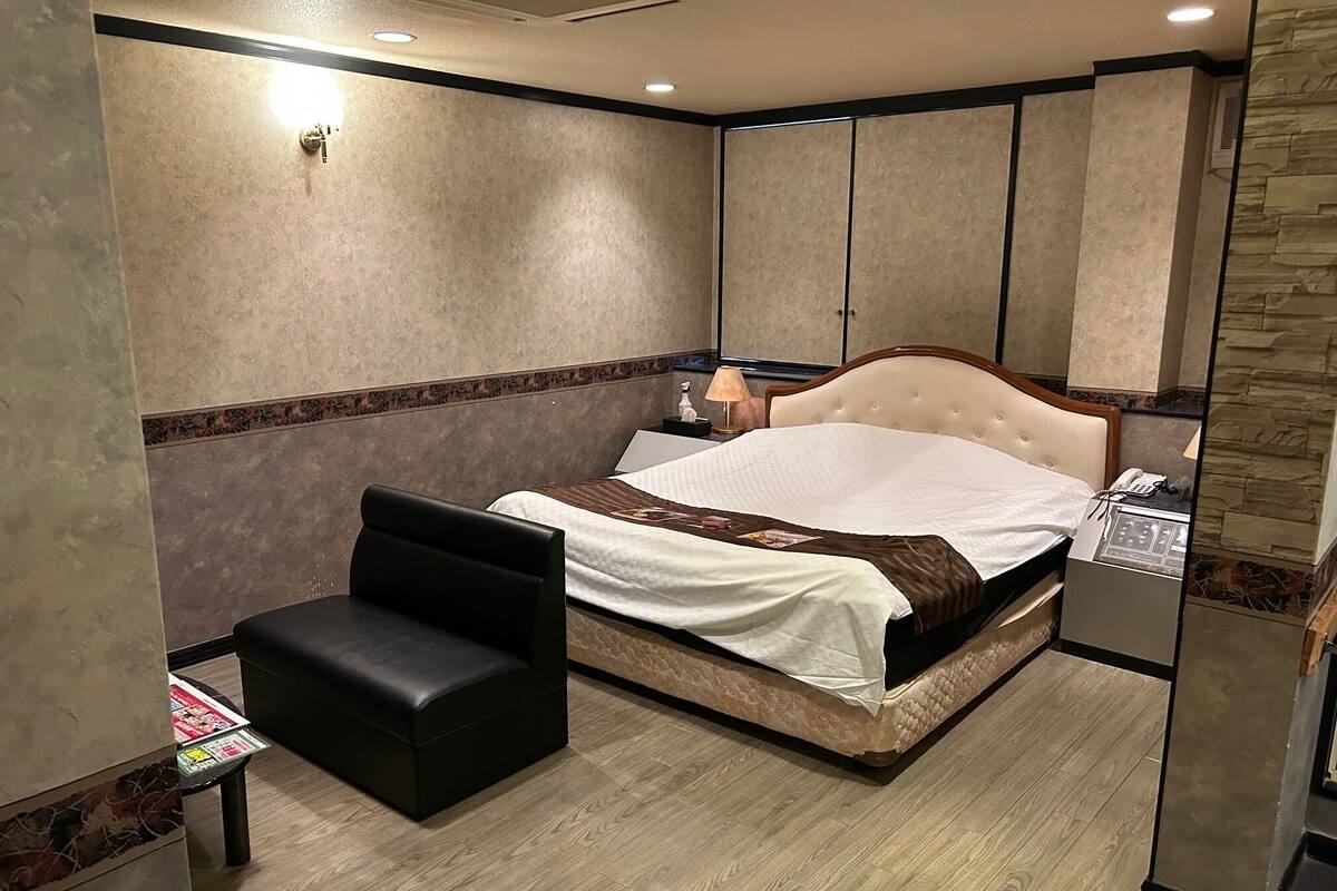 A hotel room with a bed, a small black chair, a nightstand with a lamp, and a telephone.