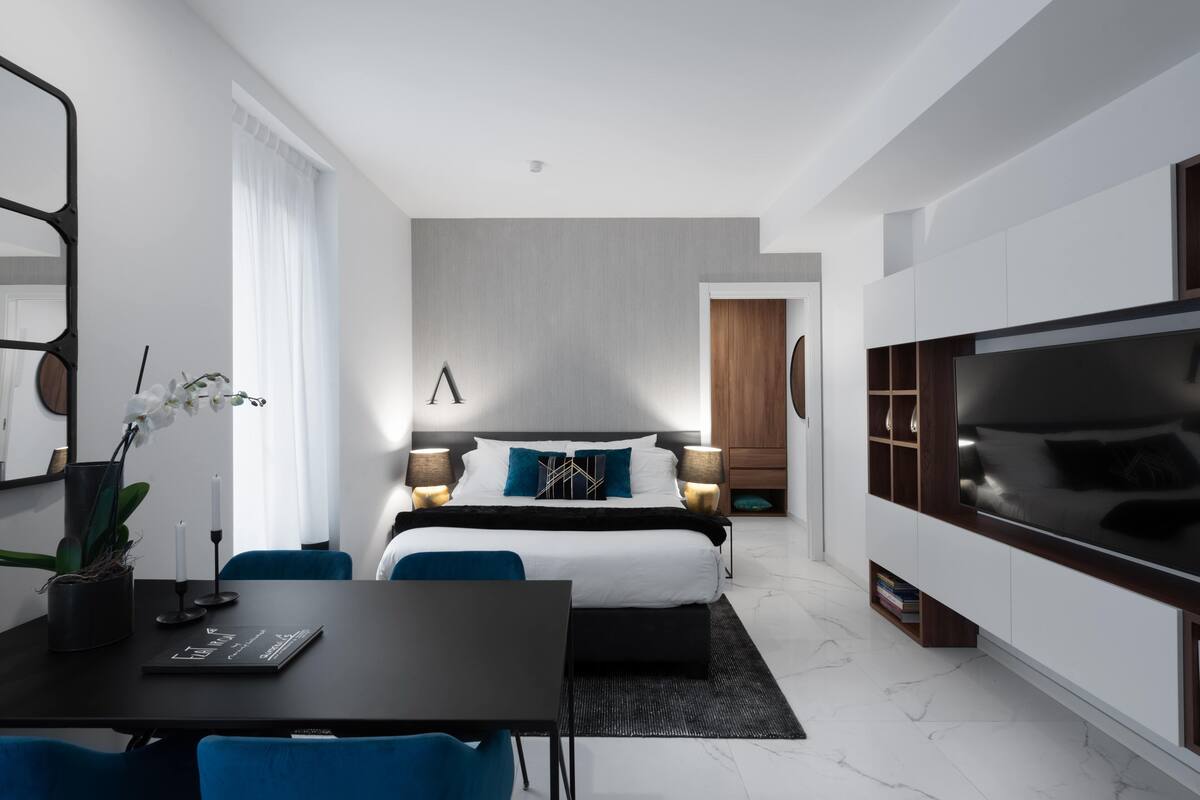 Una camera d'albergo moderna con un ampio letto, lampade ai comodini, una TV, un tavolo da pranzo e un'area salotto.