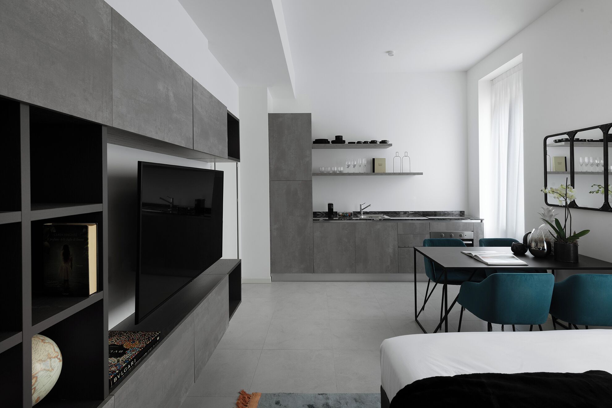 Ein modernes, minimalistisches Hotelzimmer mit einem großen Flachbildfernseher, einem Essbereich mit Tisch und Stühlen sowie einer durch eine offene Tür erkennbaren Küchenzeile.