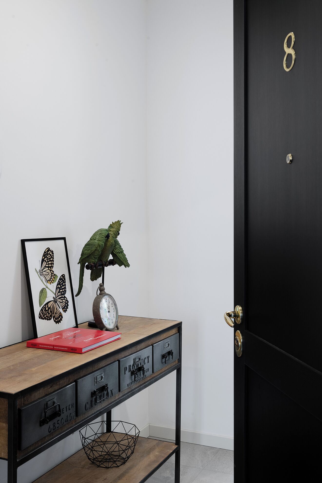 A wooden desk with drawers, a clock, a framed butterfly picture, a potted plant, and a black door with the number 8.