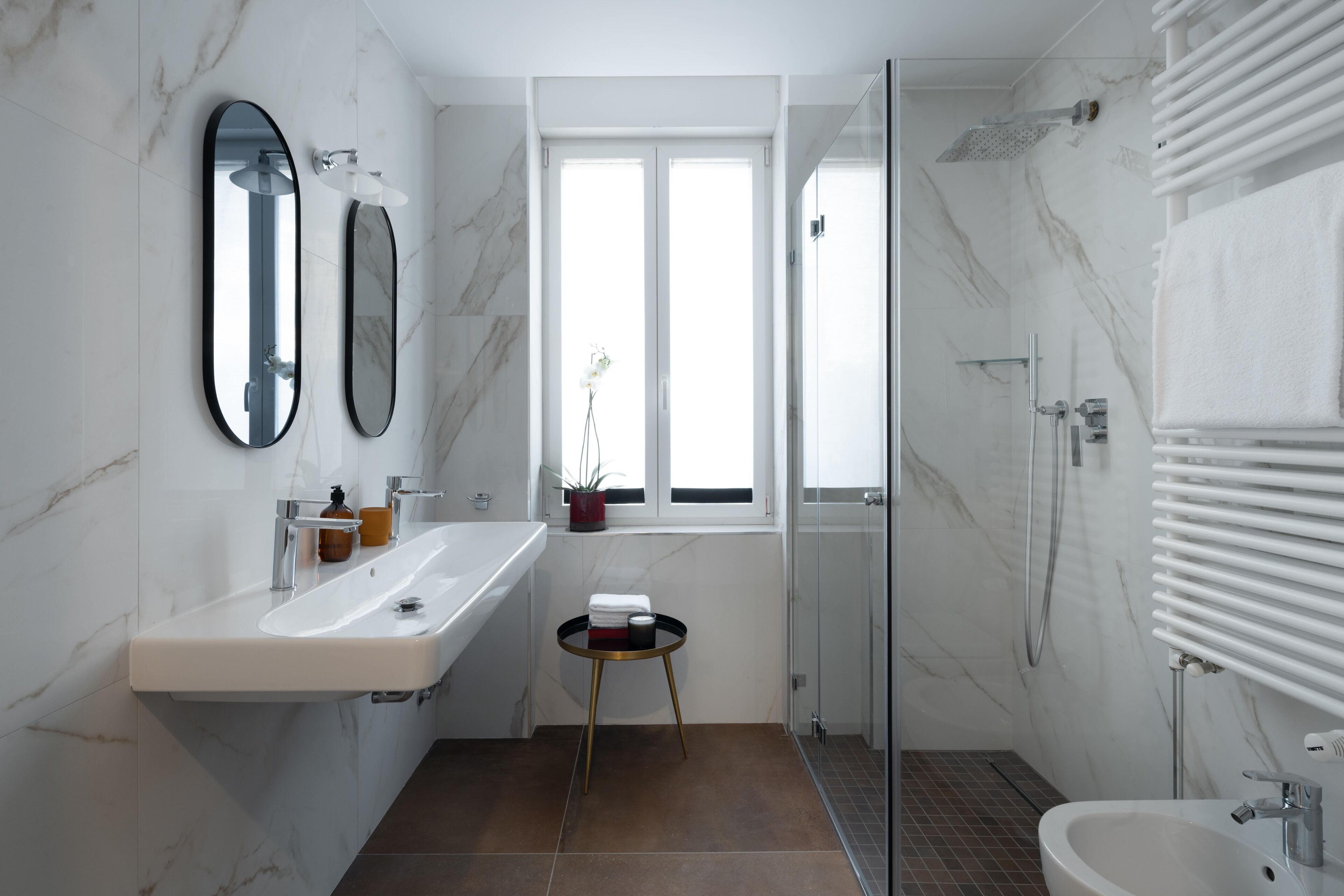 Ein modernes Badezimmer mit einem weißen Waschbecken, zwei Spiegeln, einer Duschkabine aus Glas und einem kleinen runden Tisch mit Pflegeprodukten.