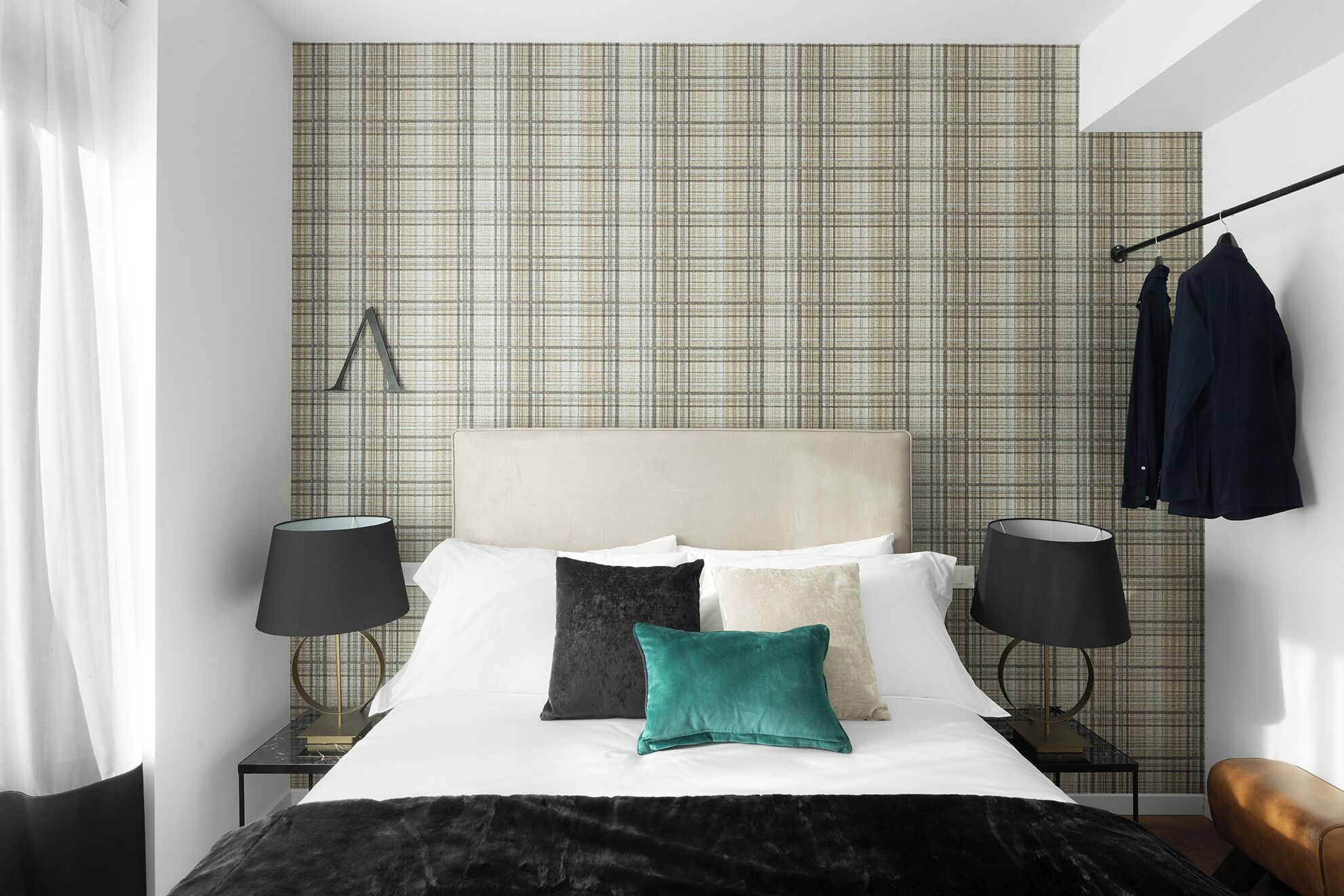A modern bedroom with a plaid wallpaper, a headboard, two bedside tables with lamps, a bed with multiple pillows, and a hanging coat rack.