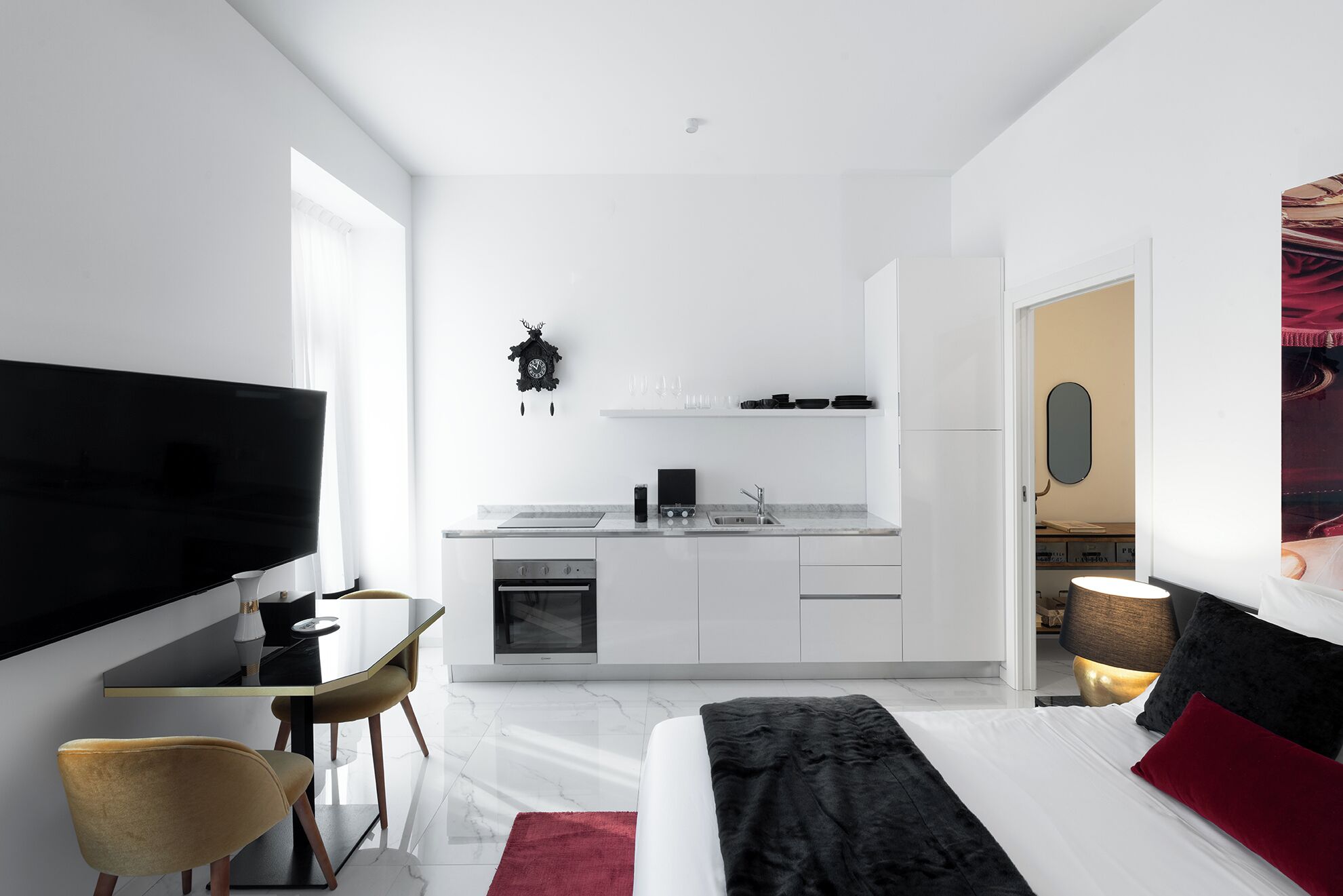 Camera d'albergo moderna con televisione a schermo piatto, zona pranzo con tavolo e sedie, angolo cottura con fornelli e lavello, e un letto con cuscino rosso.