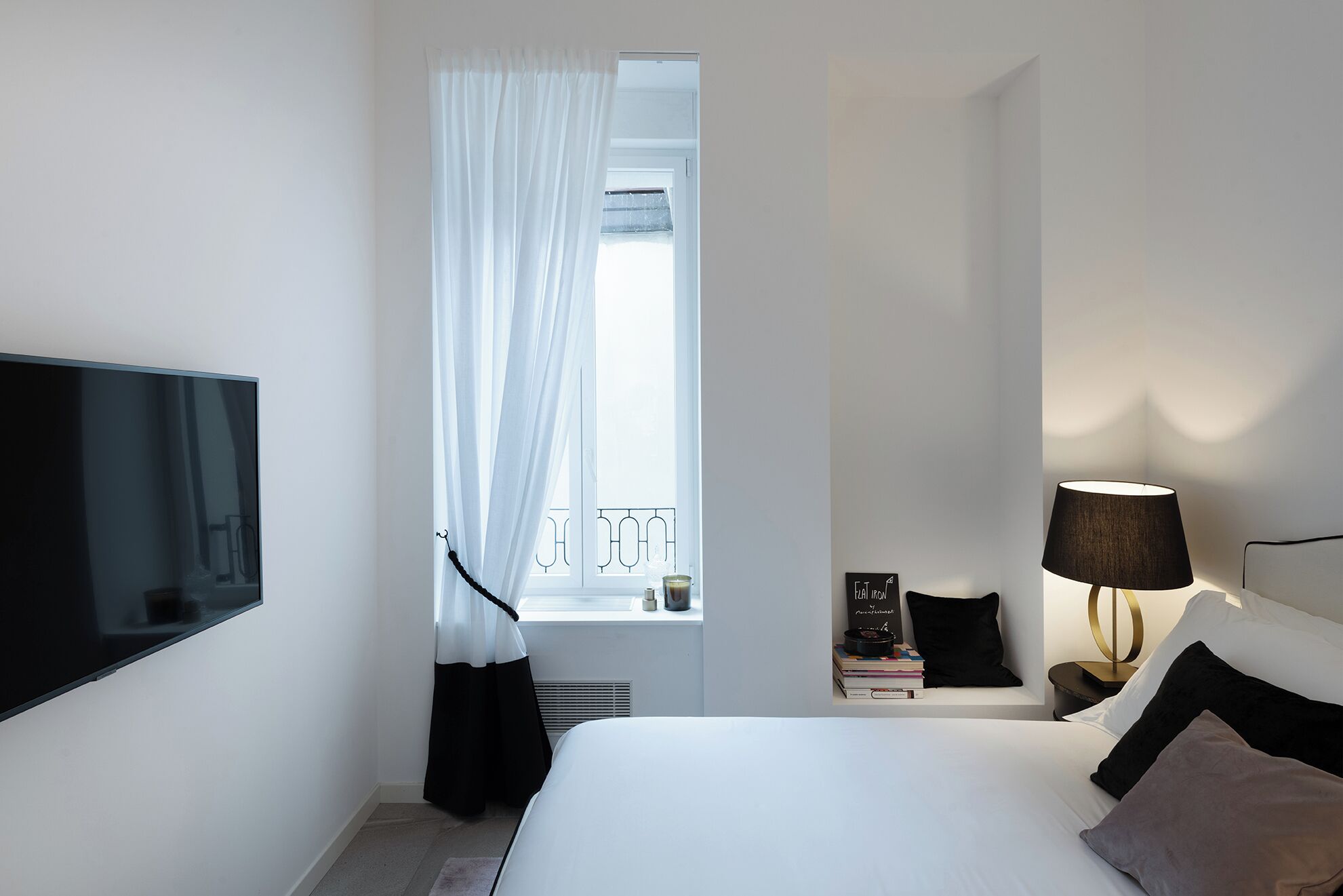 A hotel room with a bed, a TV, a bedside table with books and a lamp, a window with sheer curtains, and a view of a balcony.