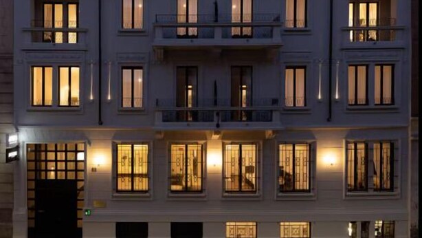 Un edificio de varios pisos con ventanas e balcones iluminados, coches aparcados al frente y un cielo despejado.