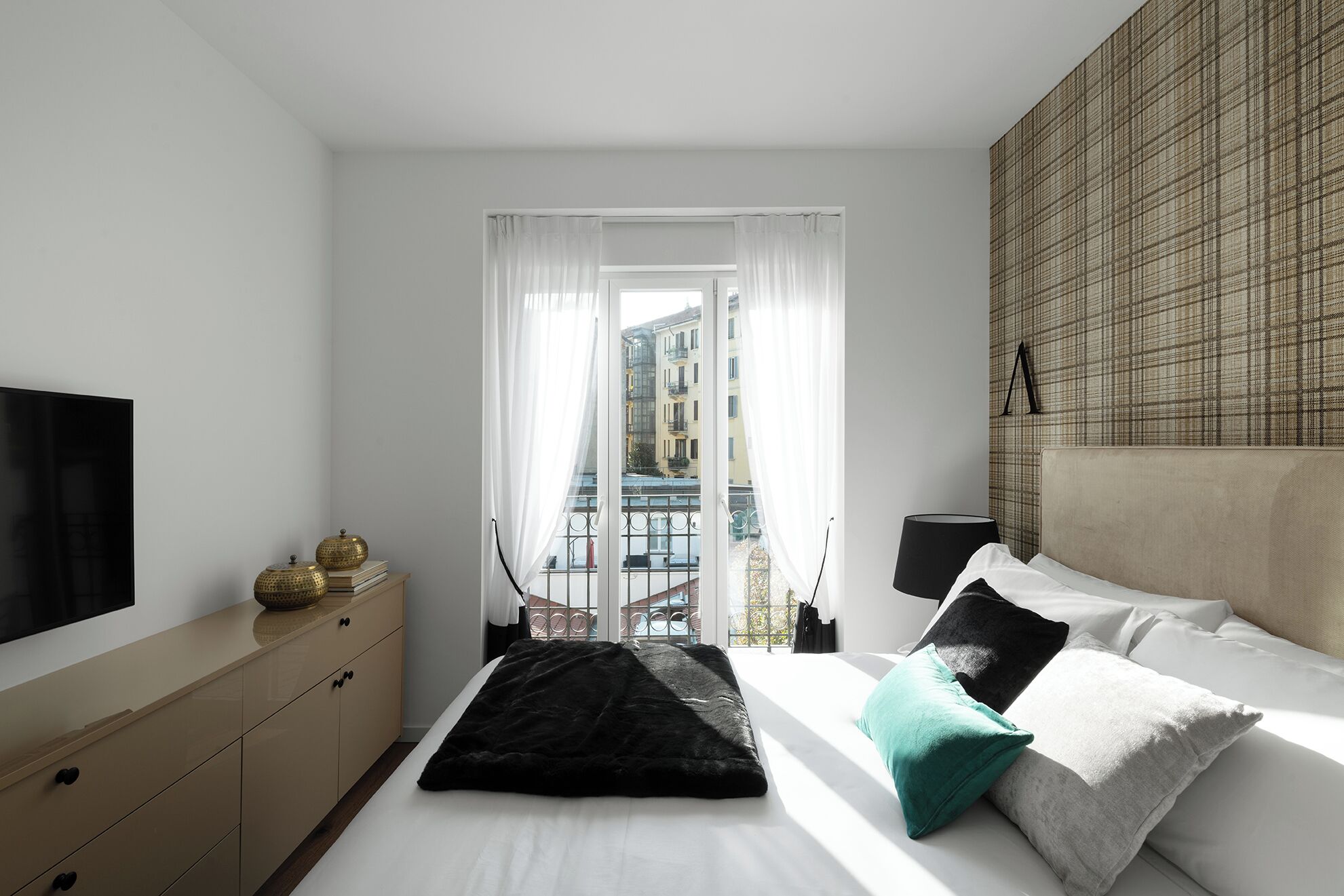 Een moderne slaapkamer met een bed, een televisie, een dressoir en uitzicht op gebouwen door dunne gordijnen.