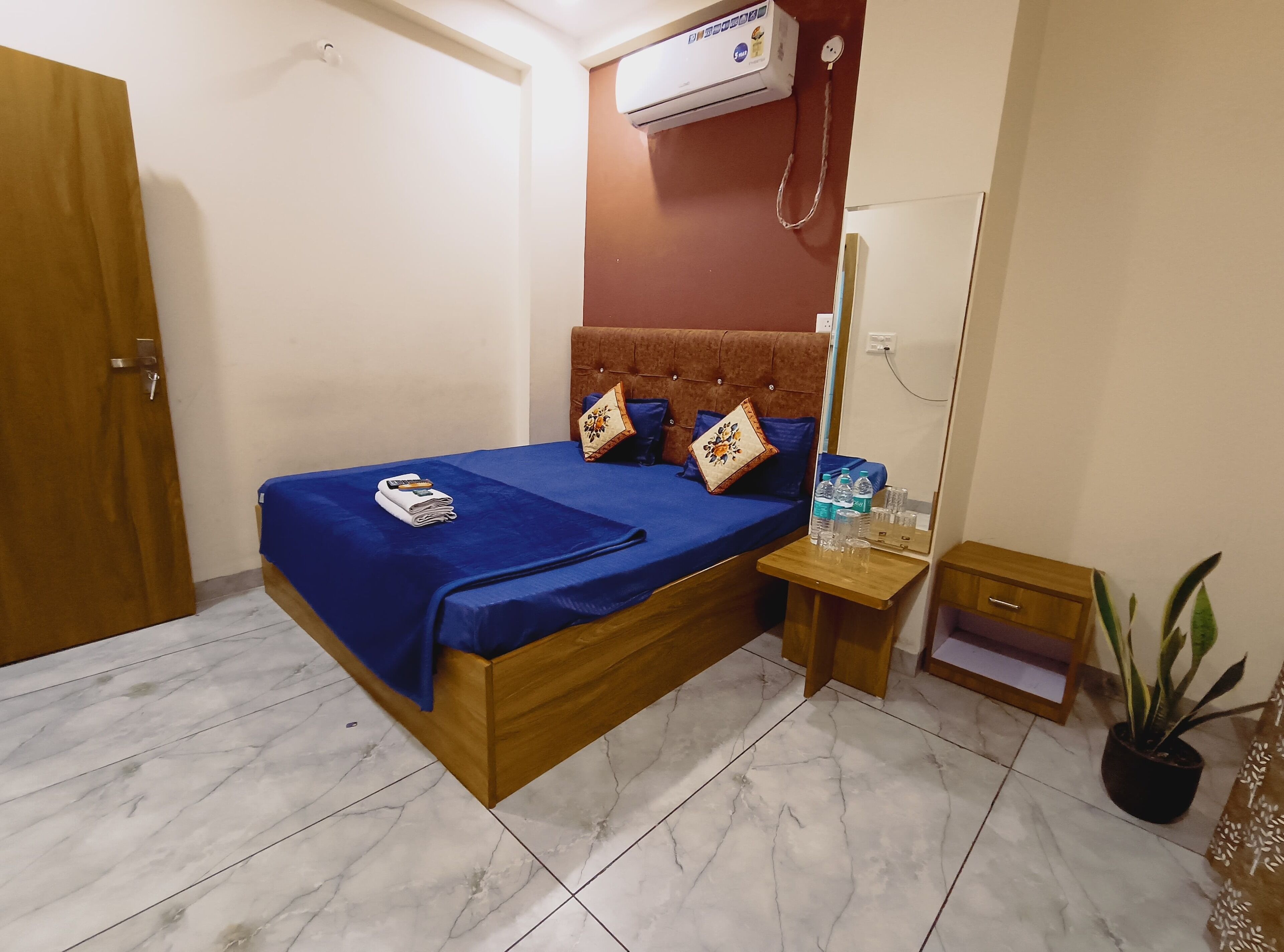 A hotel room with a bed, a nightstand, a mirror, a potted plant, and an air conditioning unit.