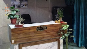 A reception desk with a marble front, wooden paneling, and a sign that reads "RECEPTION". There are potted plants on the desk and a red carpet in front. - Hotel Laxmi villa (Ujjain)
